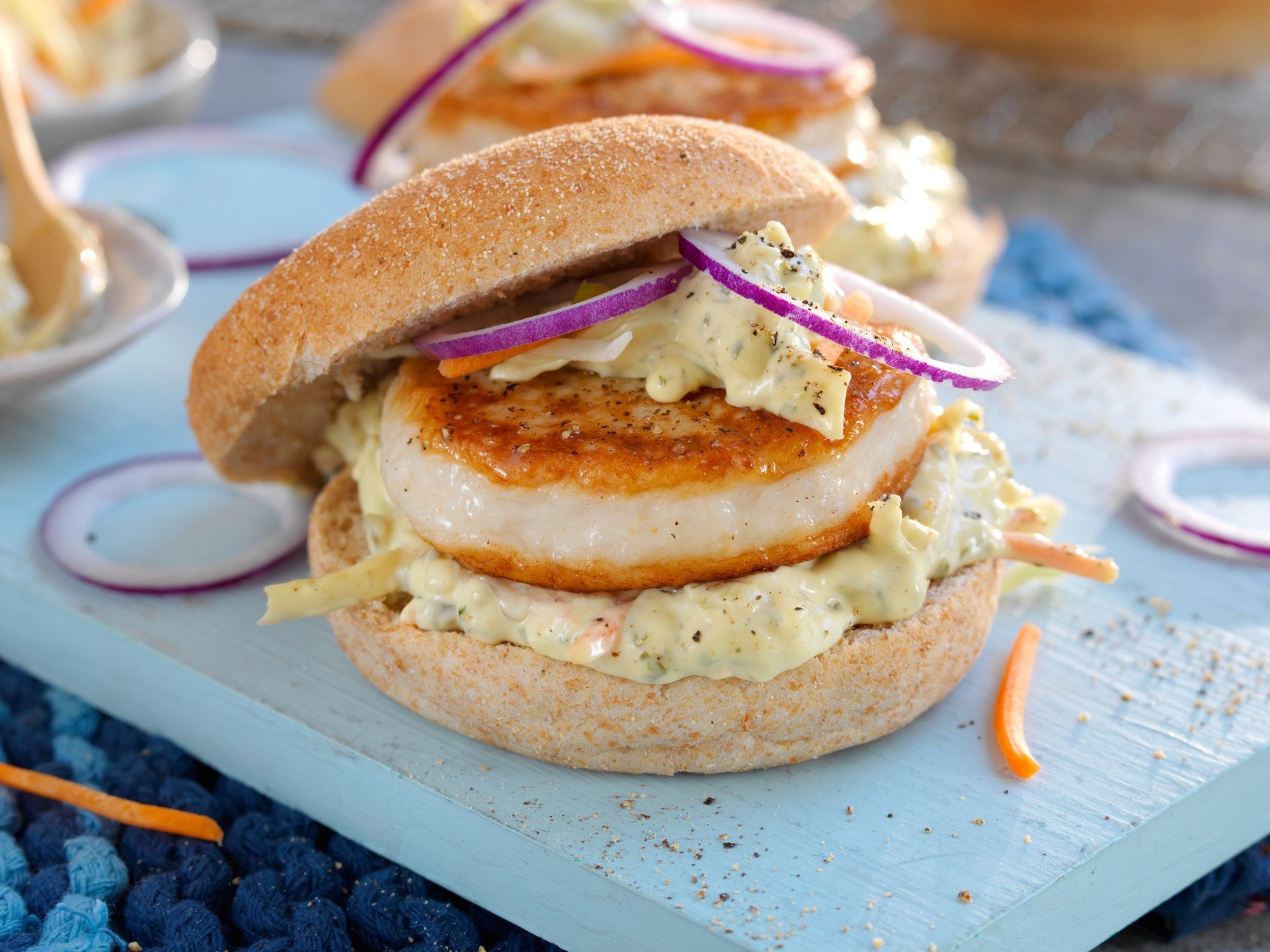 Fiskeburger med coleslaw og skiver av rødløk i et brød, servert på en lys skjæreflate.