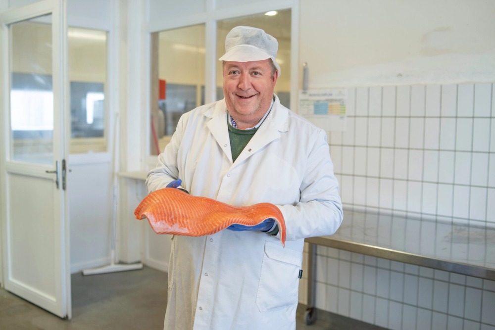Person i hvit labfrakk holder en stor laksefilet i hendene på et laboratorium eller fiskerifasilitet.