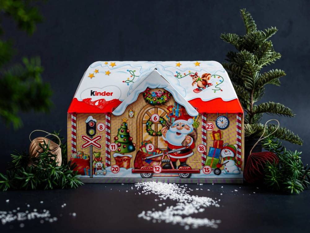 En julekalender i form av et lite hus med juledekorasjoner og en figur av julenissen foran, omgitt av granbar og julepynt.