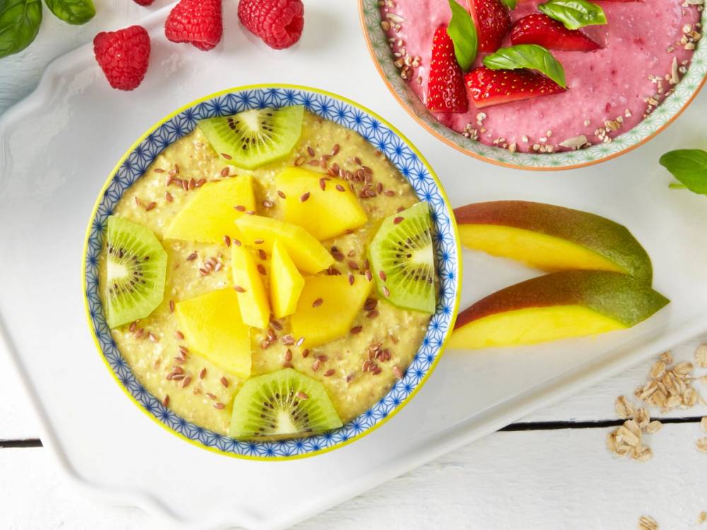 En skål med fruktsalat som inneholder mango, kiwi og ananas, omgitt av flere fruktstykker og bær på et hvitt bord.