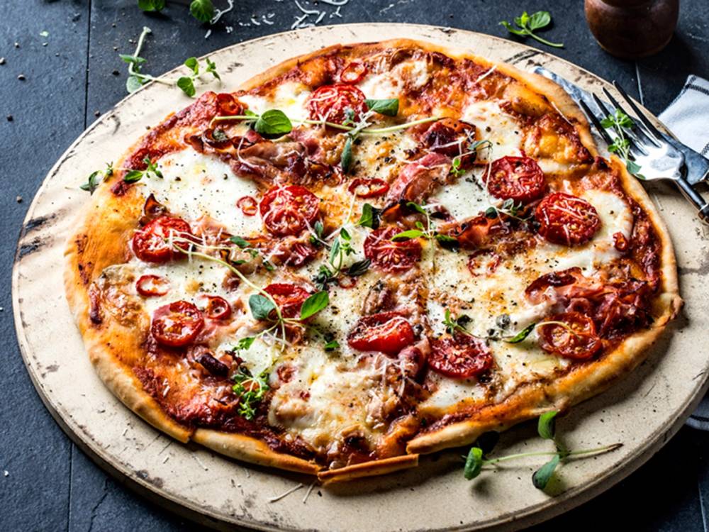 Hjemmelaget italiensk pizza med bacon, tomatskiver og frisk basilikum på en treplate.