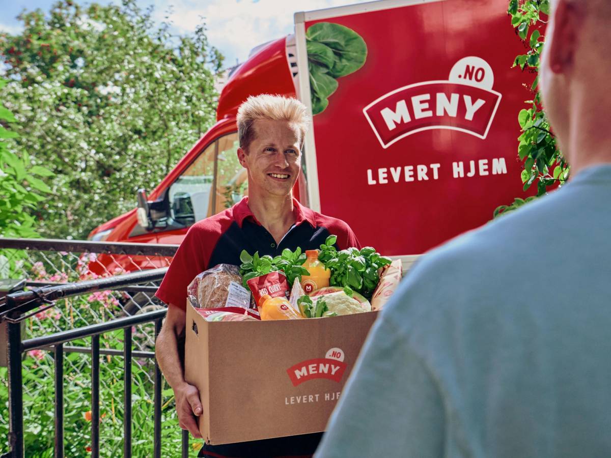 En person leverer en matkurv med dagligvarer til en kunde utendørs, med en rød leveringsbil i bakgrunnen.