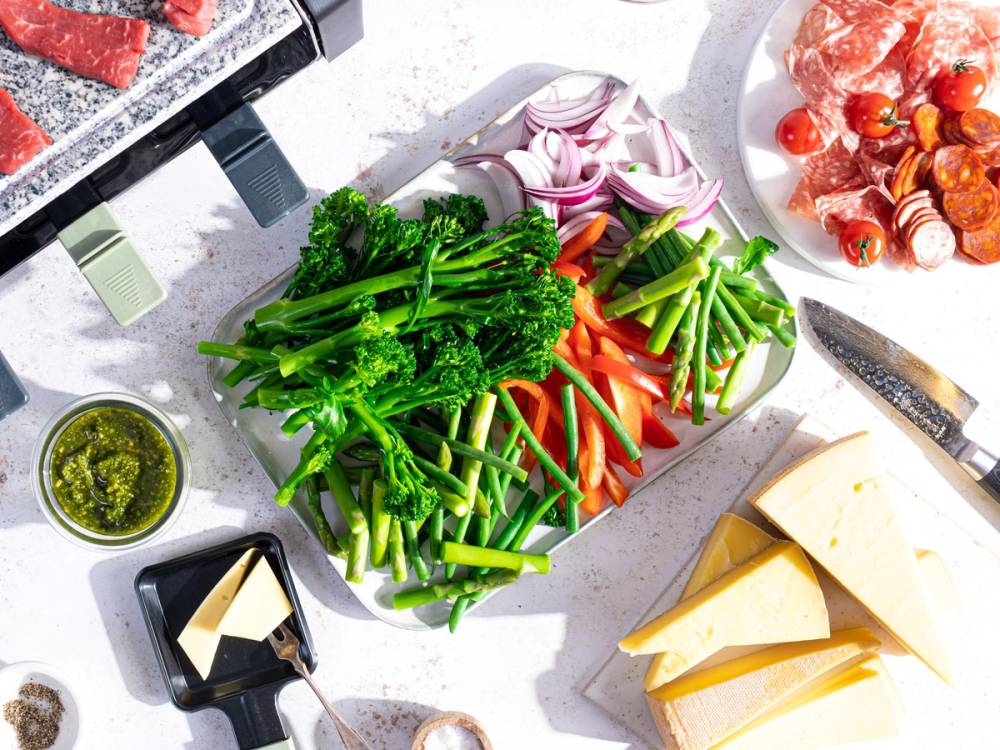 Skjærebrett med ferske grønnsaker som brokkoli, paprika og agurk, omgitt av ost, skinke og tilbehør til raclette.