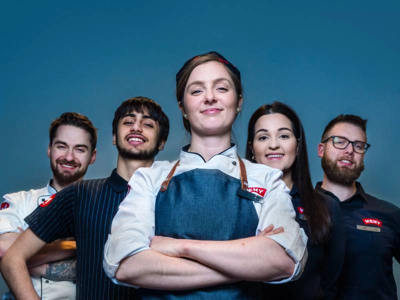 En gruppe personer i uniformer står sammen med armene i kors mot en blå bakgrunn, med den ene personen i midten i fokus.