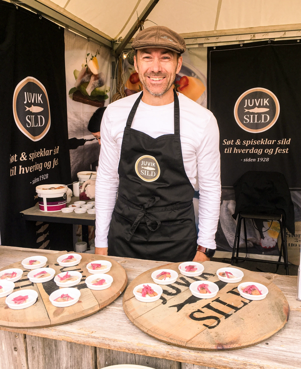 Person med forklær foran en bord med små tallerkener med mat, i en utendørs stand med Juvik Sild-logoer i bakgrunnen.