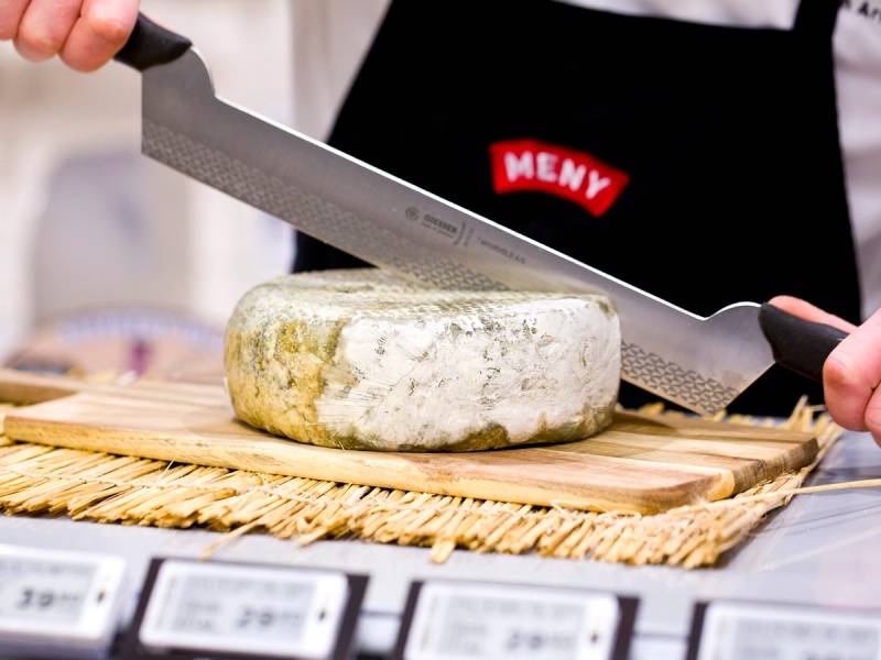 Person skjærer en rund ost med en stor kniv på et skjærebrett, med en person iført svart forkle med rødt merke i bakgrunnen.