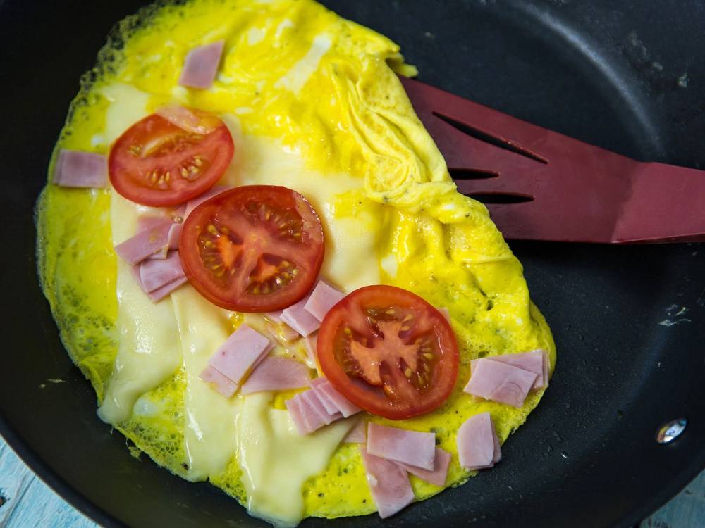 Omelett med skiver av tomat og små biter av skinke i en stekepanne, med en rosa stekespade ved siden av.