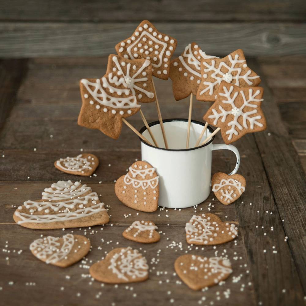 Pepperkakefigurer med hvit glasur pyntet på en treplate, noen ligger på bordet rundt en kopp med pepperkakefigurer på pinne.