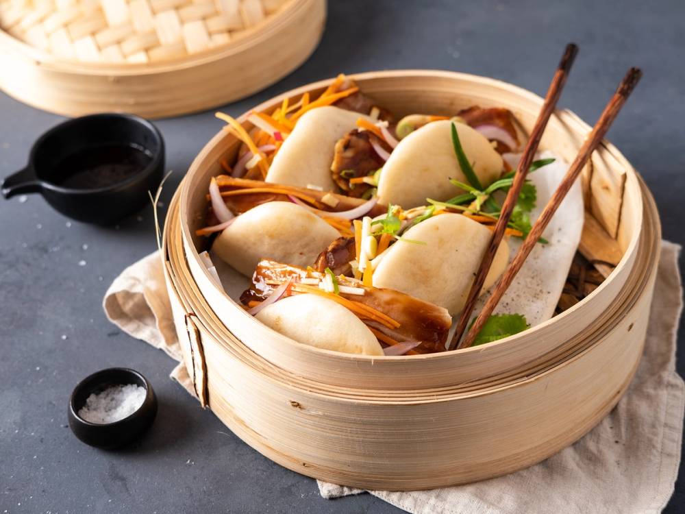 Steamed buns med kjøtt og grønnsaker i en bambuskurv, omgitt av små skåler med saus.