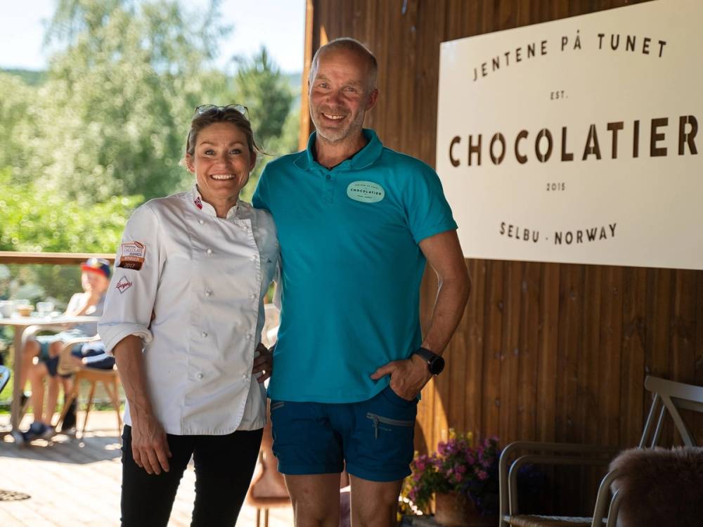 To kvinner står sammen under et tak, med en plakat som sier 'Jentene på Tunet' og 'Chocolatier' i bakgrunnen, på en utendørs arena i Norge.