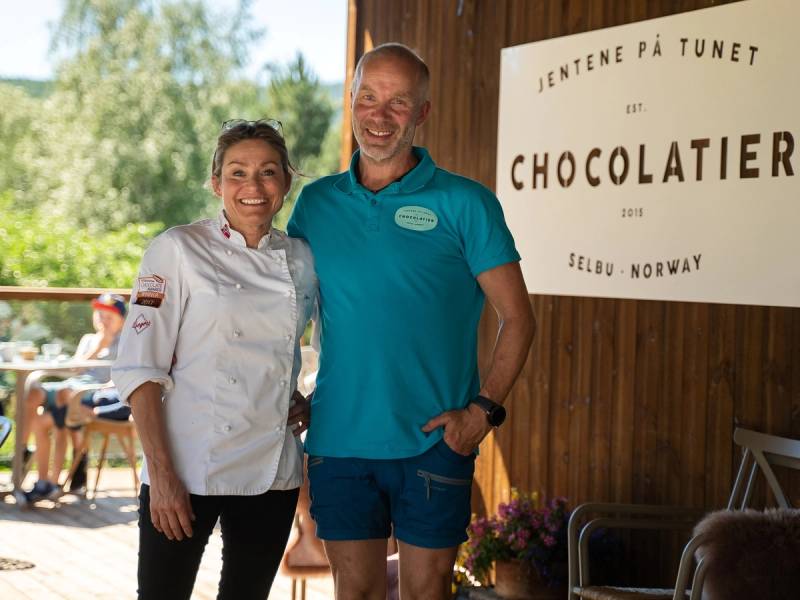 To kvinner står sammen under et tak, med en plakat som sier 'Jentene på Tunet' og 'Chocolatier' i bakgrunnen, på en utendørs arena i Norge.