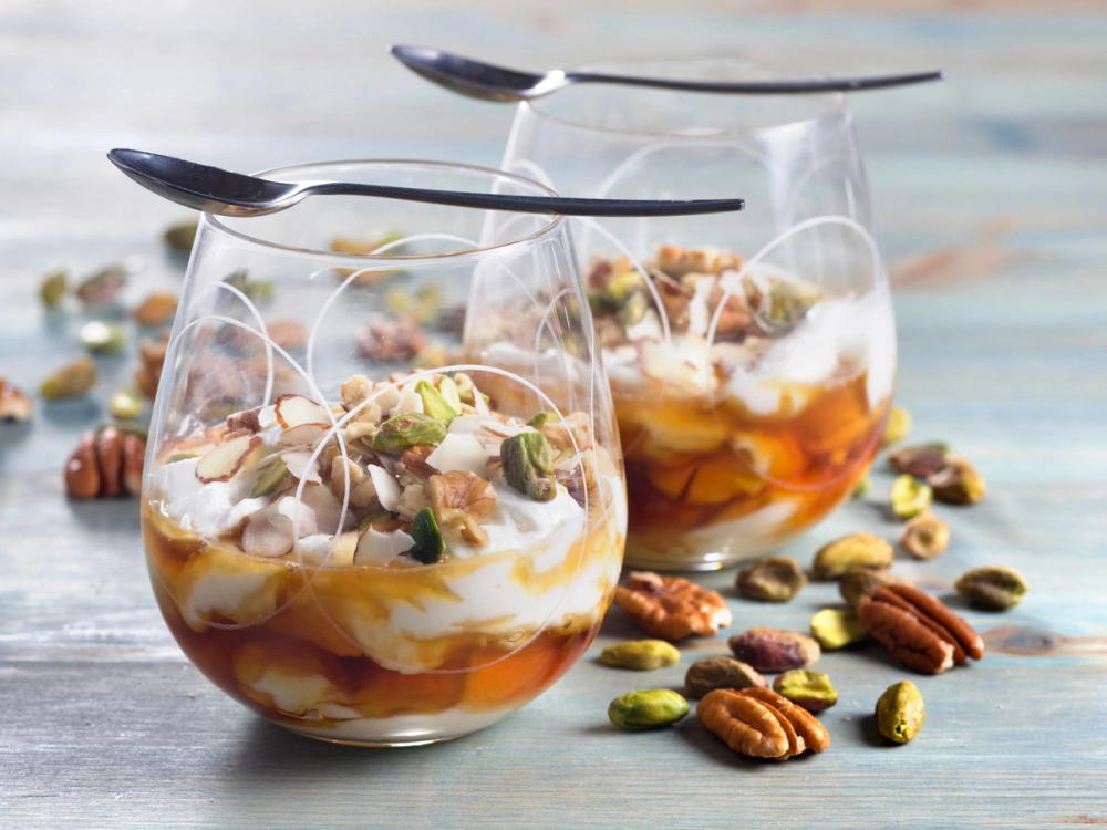 To glass med yoghurt med honning og nøtter, omgitt av spredte nøtter på bordet.
