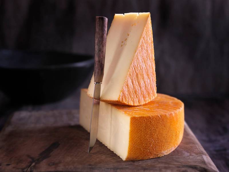 Ulike typer ost, inkludert en blokk og en skive, på et trebrett med en ostehøvel ved siden av.