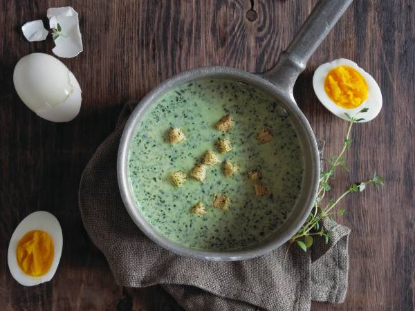 Spinatsuppe med egg i en gryte på et bord, omgitt av kokte egg og en liten plante.