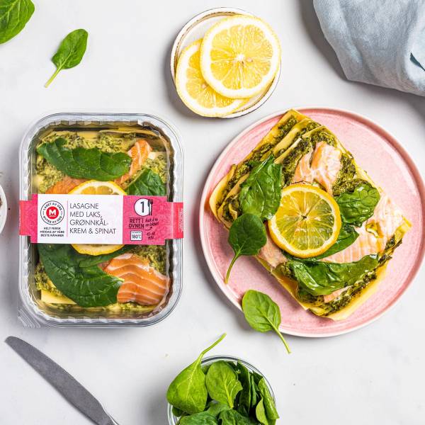 Ferdiglaget lakse-lasagne med grønnsaker og sitron på en rosa tallerken, omgitt av sitronbiter og ferske spinatblader.