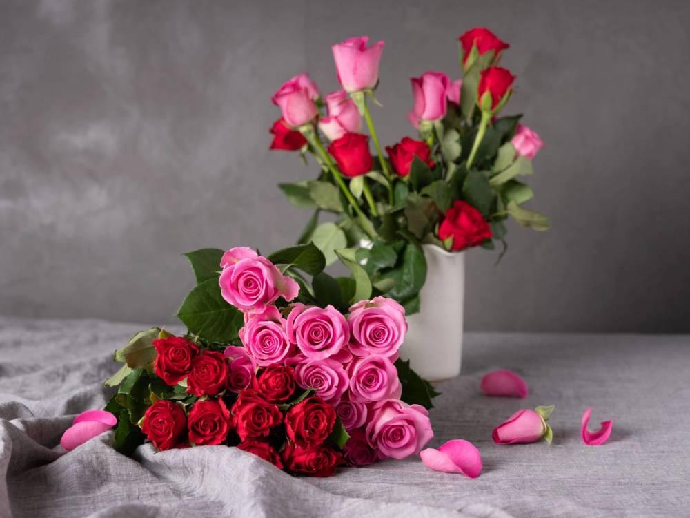 En bukett med rosa og røde roser plassert på et bord med noen spredte rosenblader rundt.