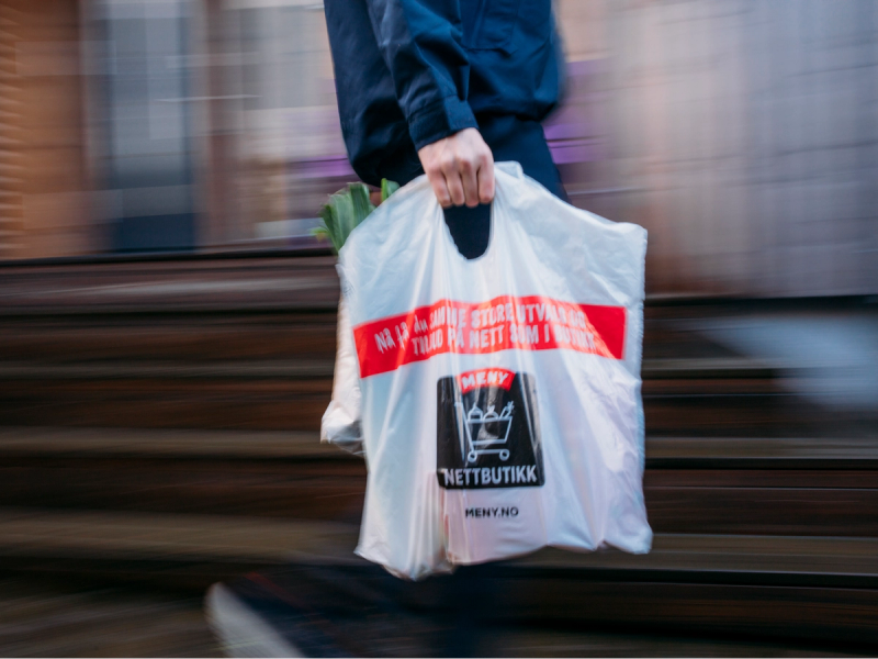 Person som bærer en plastpose med nettbutikkens logo og tekst, mens de går langs en jernbanespor eller togskinner.