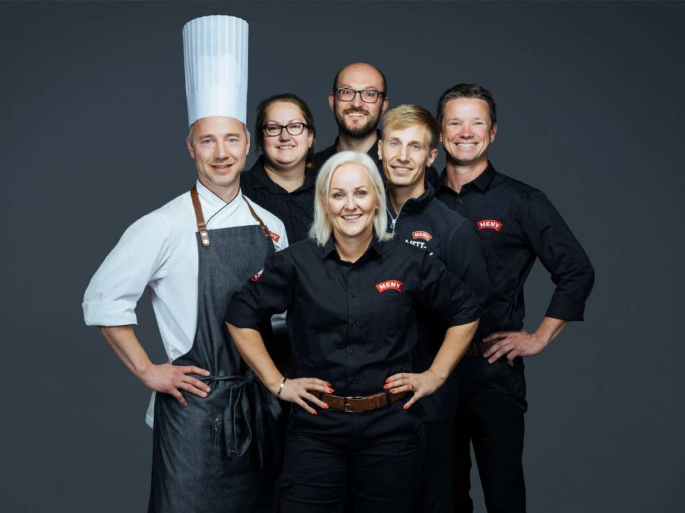 En gruppe personer, inkludert en kok med hvit kokkehatt, står sammen i et studio med mørk bakgrunn.