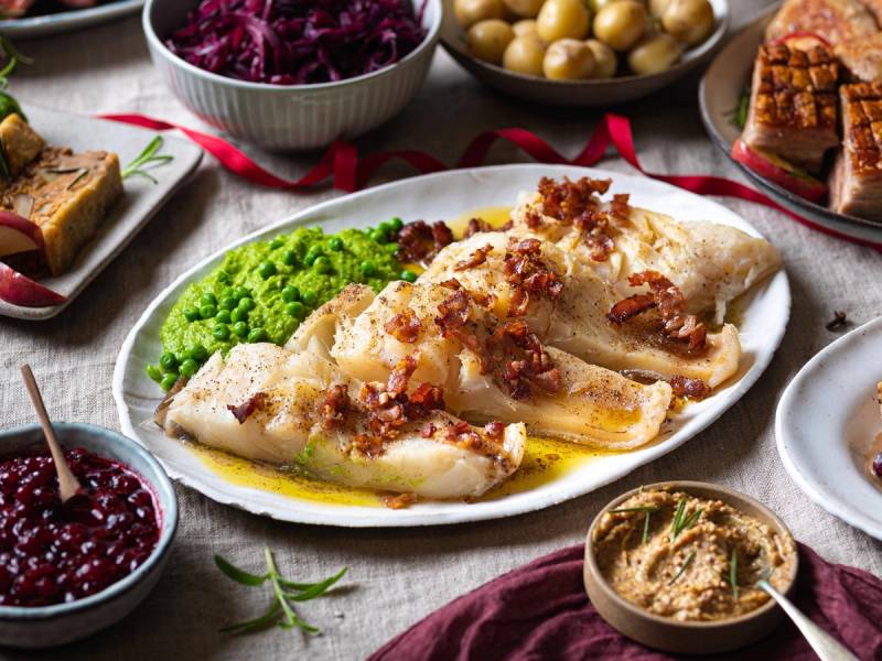 Stekt torsk med bacon og grønnsaker på et serveringsfat, omgitt av ulike småretter og tilbehør på bordet.