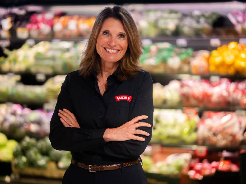 En kvinne står med armene krysset i en matbutikk, iført en svart skjorte med MENY-logo, omgitt av frukt og grønnsaker.