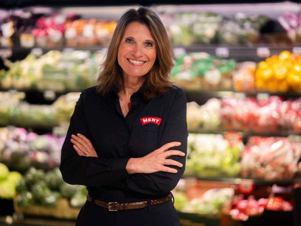 En kvinne står med armene krysset i en matbutikk, iført en svart skjorte med MENY-logo, omgitt av frukt og grønnsaker.