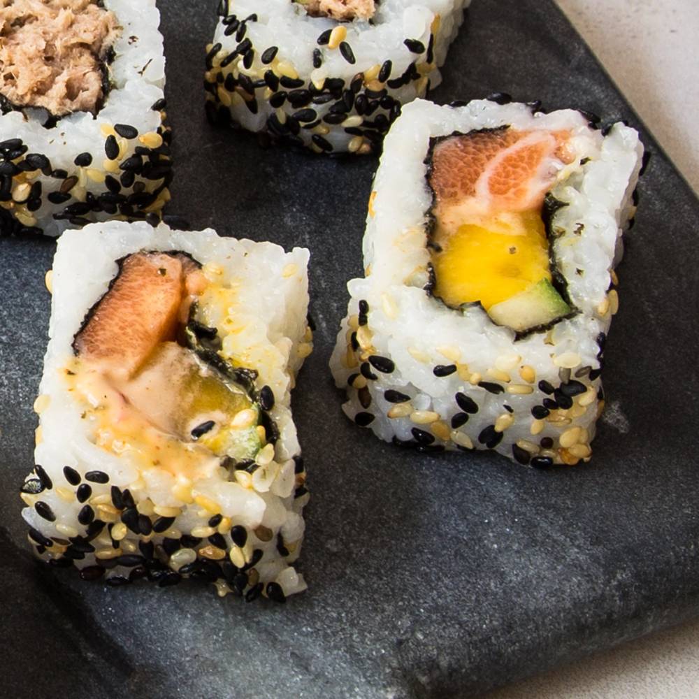 Sushi maki med laks, agurk og egg, pyntet med svarte sesamfrø på en svart steinplate.