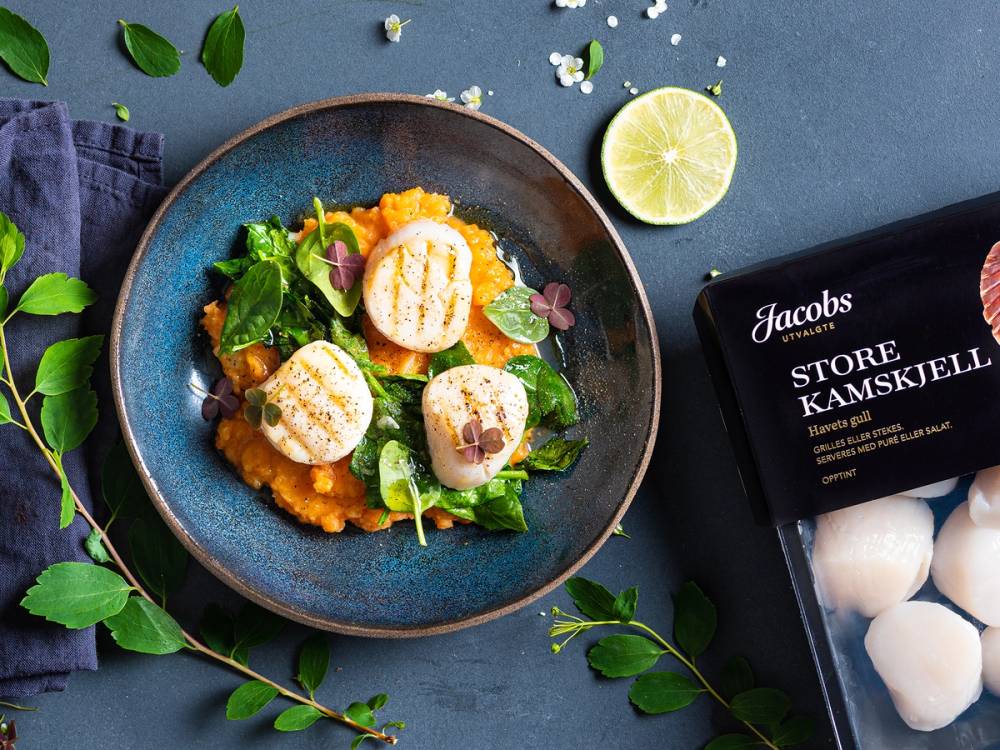 Kamskjell servert på en tallerken med grønnsaker og en sitronhalvdel ved siden av, med en pakke Jacobs utvalgte kamskjell i bakgrunnen.