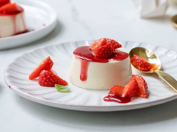 Panna cotta med gløggmarinerte jordbær på en hvit tallerken med en gullskje ved siden av.