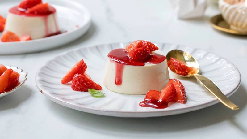 Panna cotta med gløggmarinerte jordbær på en hvit tallerken med en gullskje ved siden av.