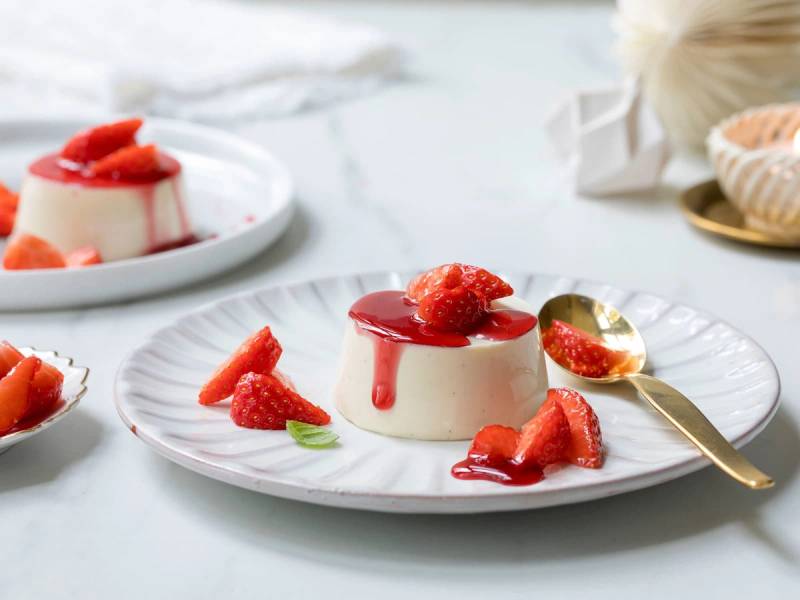 Panna cotta med gløggmarinerte jordbær på en hvit tallerken med en gullskje ved siden av.