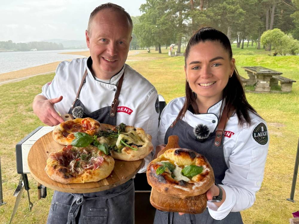 To personer holder opp pizzaer under åpen himmel med grøntområde og vann i bakgrunnen.