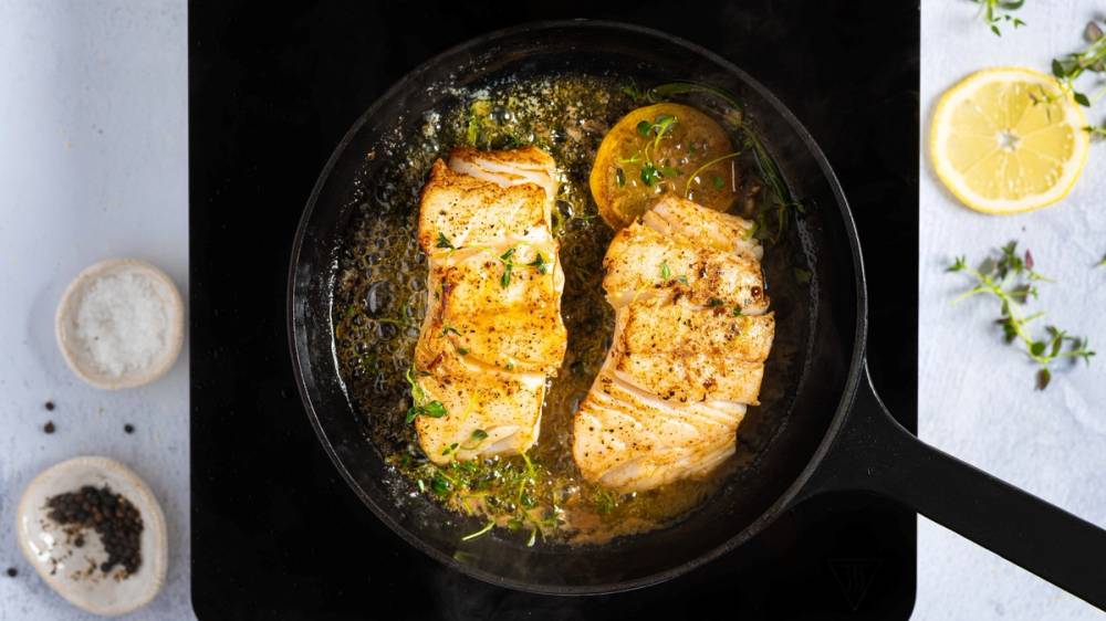 Steker torsk i en panne med sitron og urter, omgitt av sitronbåter og krydder på bordet.