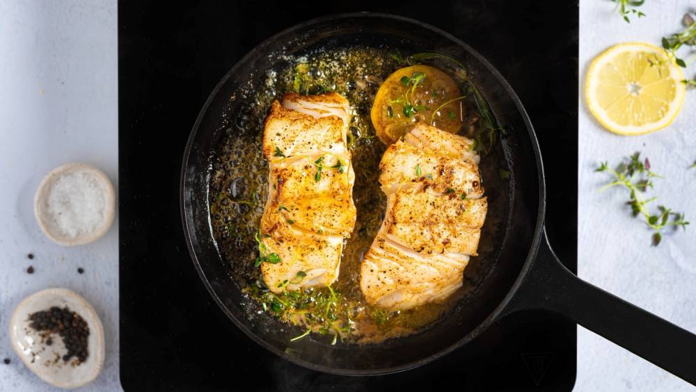 Steker torsk i en panne med sitron og urter, omgitt av sitronbåter og krydder på bordet.
