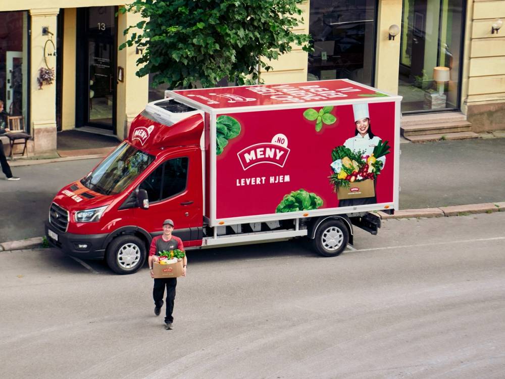 En mann står foran en rød leveringsbil med Meny-logo og bilder av grønnsaker og en person som holder en kurv med mat, parkert foran en butikk eller bygning.