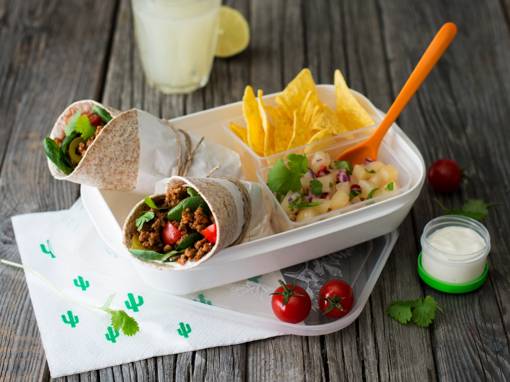 Tacorester med to wraps, chips, salat, og en liten skål med dip på et bord med tomater og en limebåt.