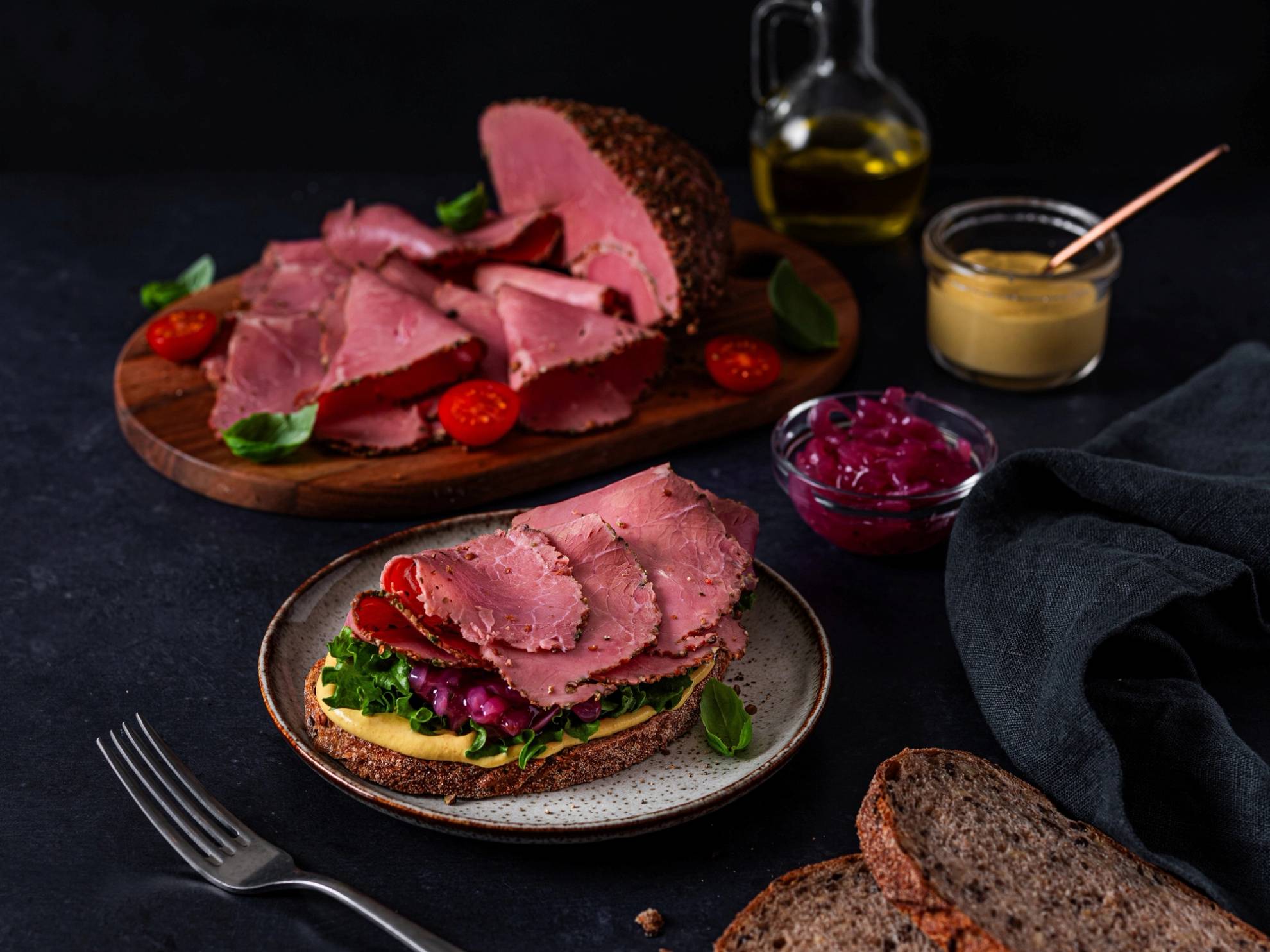 Skinke og kjøttpålegg på en tallerken med salat og brød, omgitt av pålegg og sauser på et mørkt bord.