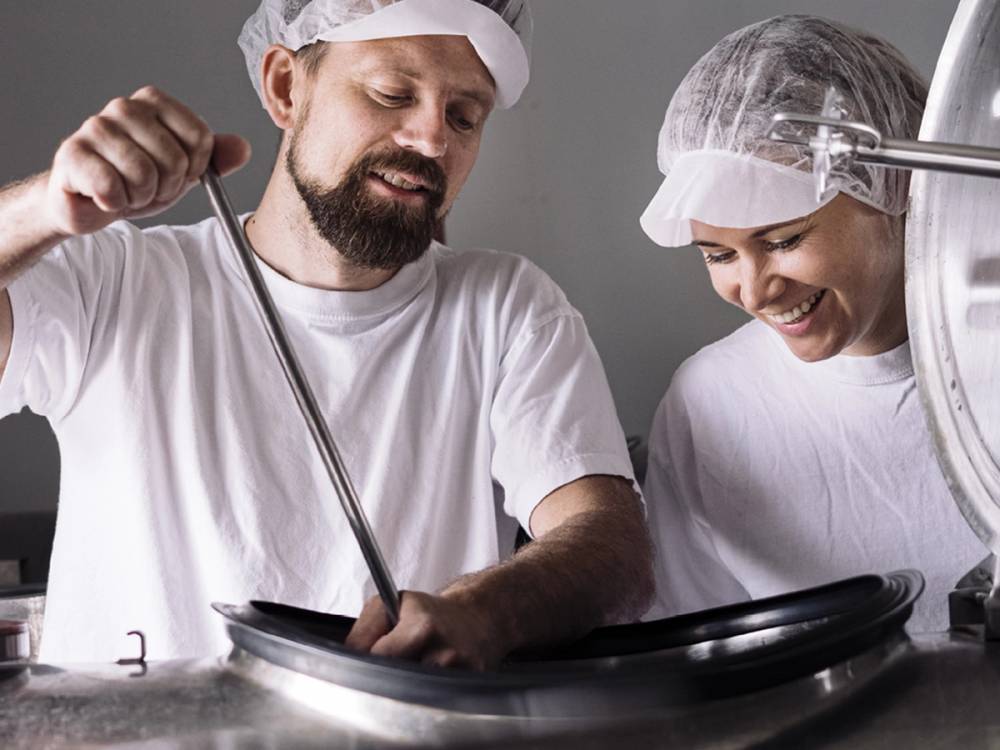 To personer lager iskrem i en iskremfabrikk, med en melketank i bakgrunnen.