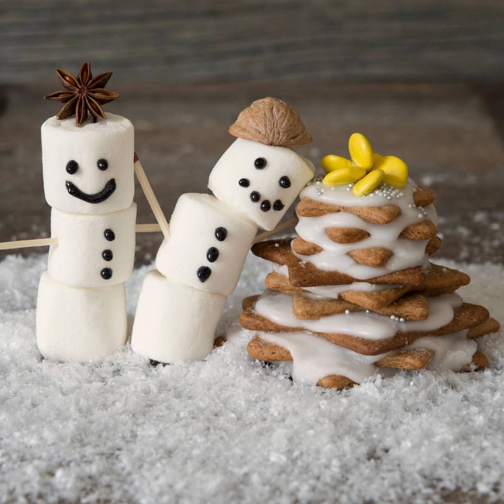 Barn lager pepperkakefigurer med marshmallows og pynt på et snødekt underlag.
