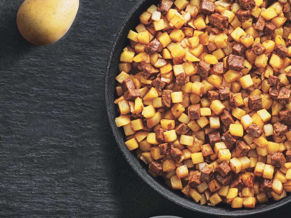 Vegetarisk pytt i panne med små terninger av grønnsaker i en svart stekepanne, med et egg ved siden av på en mørk bakgrunn.