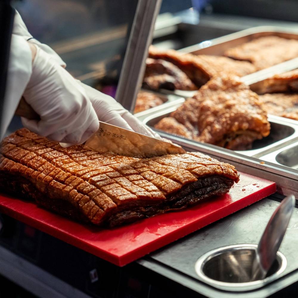 En person skjærer opp en stor, grillet kjøttstykke på en rød skjærefjøl i en matbod.