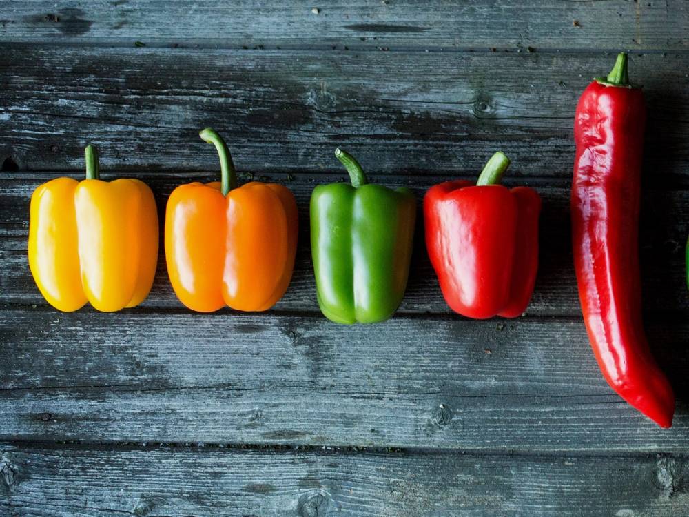 Fire paprika i gul, oransje, grønn og rød farge, plassert på en treplate, med en lang rød chili ved siden av.