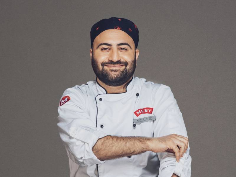 En mann i kokkeuniform med navneskilt som sier 'HENRY', står mot en grå bakgrunn, med armene krysset.