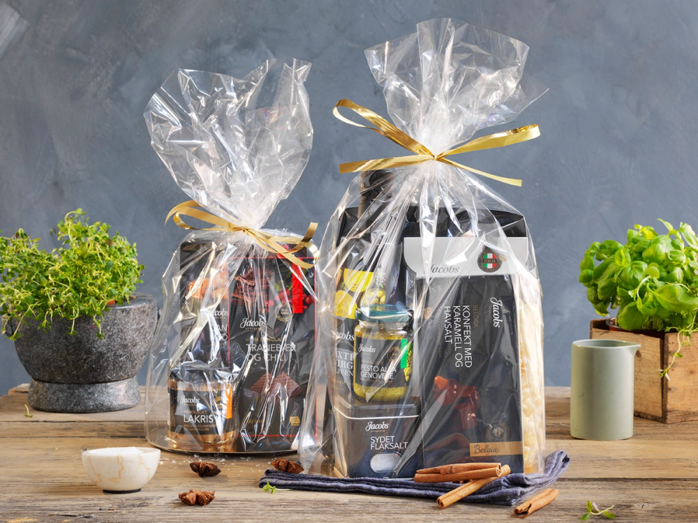 To julegaver pakket inn i gjennomsiktig plast med gull- og rød bånd, omgitt av krydder og urter på et bord.