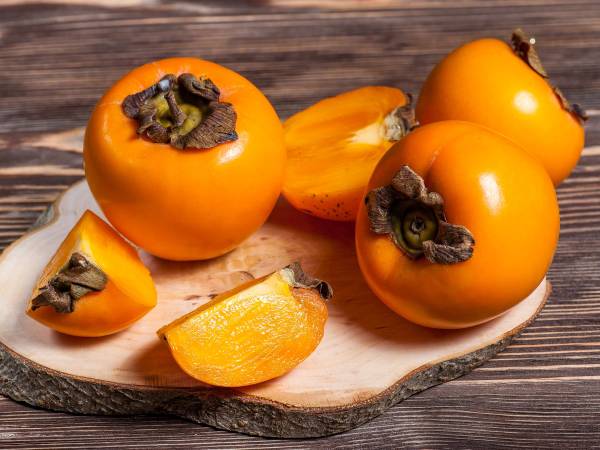 Fire persimmonfrukter, hvorav to er hele og to er delt i biter, plassert på et rundt trestykke på en trebenet bordplate.