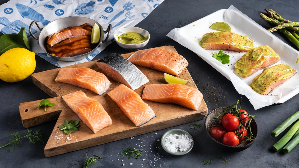 Ferske laksefileter på skjærebrett med sitron, grønnsaker og krydderurter rundt om.