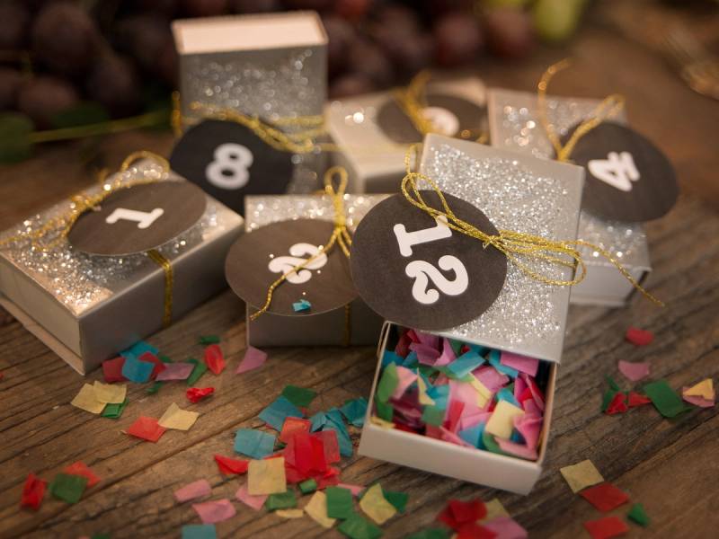 Små gaveesker med tall og glitter, omgitt av konfetti på et bord, brukt til en festkalender eller adventskalender.