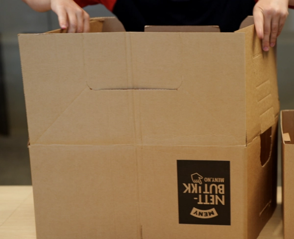 To personer holder på å pakke ut en stor pappeske med etiketten 'Nettbutikk' på siden.