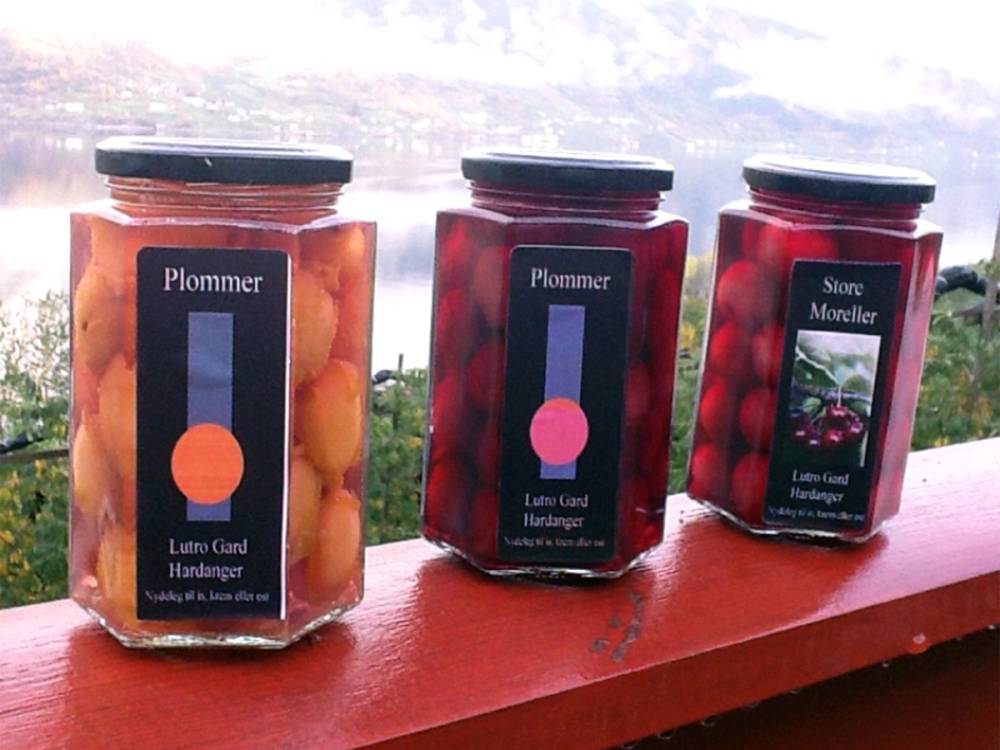 Tre glass med syltetøy eller gelé på en rød rekkverk med utsikt over vann og fjell i bakgrunnen.