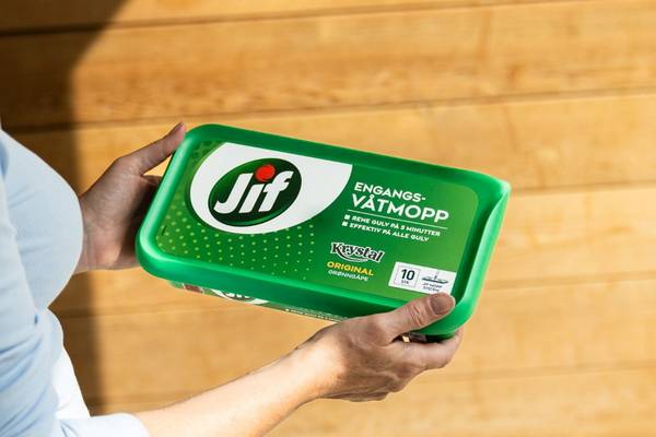 En person holder en grønn plastboks med Jif engangsvåtmopp, plassert over en trebenke, på et treverkgulv.