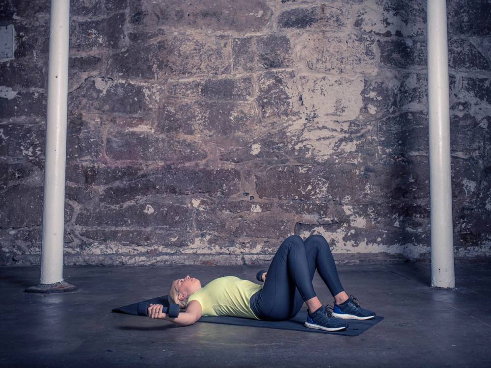En person ligger på ryggen på gulvet med en treningsmatte, iført en lys grønn topp og mørke bukser, foran en murvegg og to hvite støttestenger.